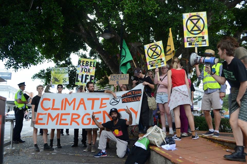Photograph of a crowd of activists gathered under a tree, holding placards and banners flanked by police. The main banner reads: “Climate Emergency”\`.