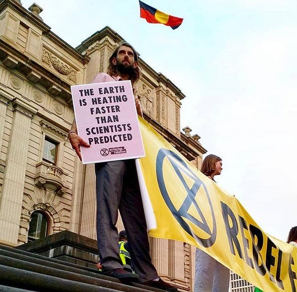 Low angle photograph of an activist standing on parliament steps, holding a placard that reads: "The earth is heating faster than scientists predicted". The indigenous flag flies in the background.